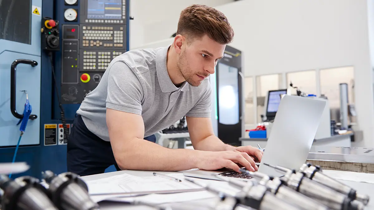 Technical Documentation for Industrial Equipment—Creation and Updates An engineer is reviewing the technical documentation for a piece of industrial equipment on a laptop, with technical drawings on the table and a CNC machine in the background